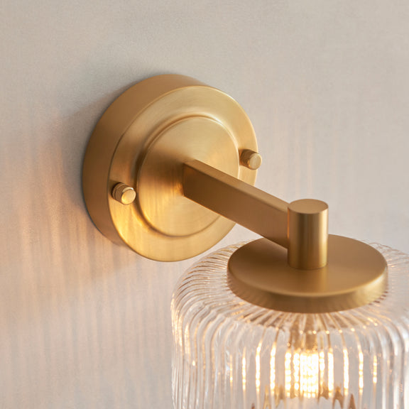 Gold wall-mounted light fixture with clear glass shade on a neutral background
