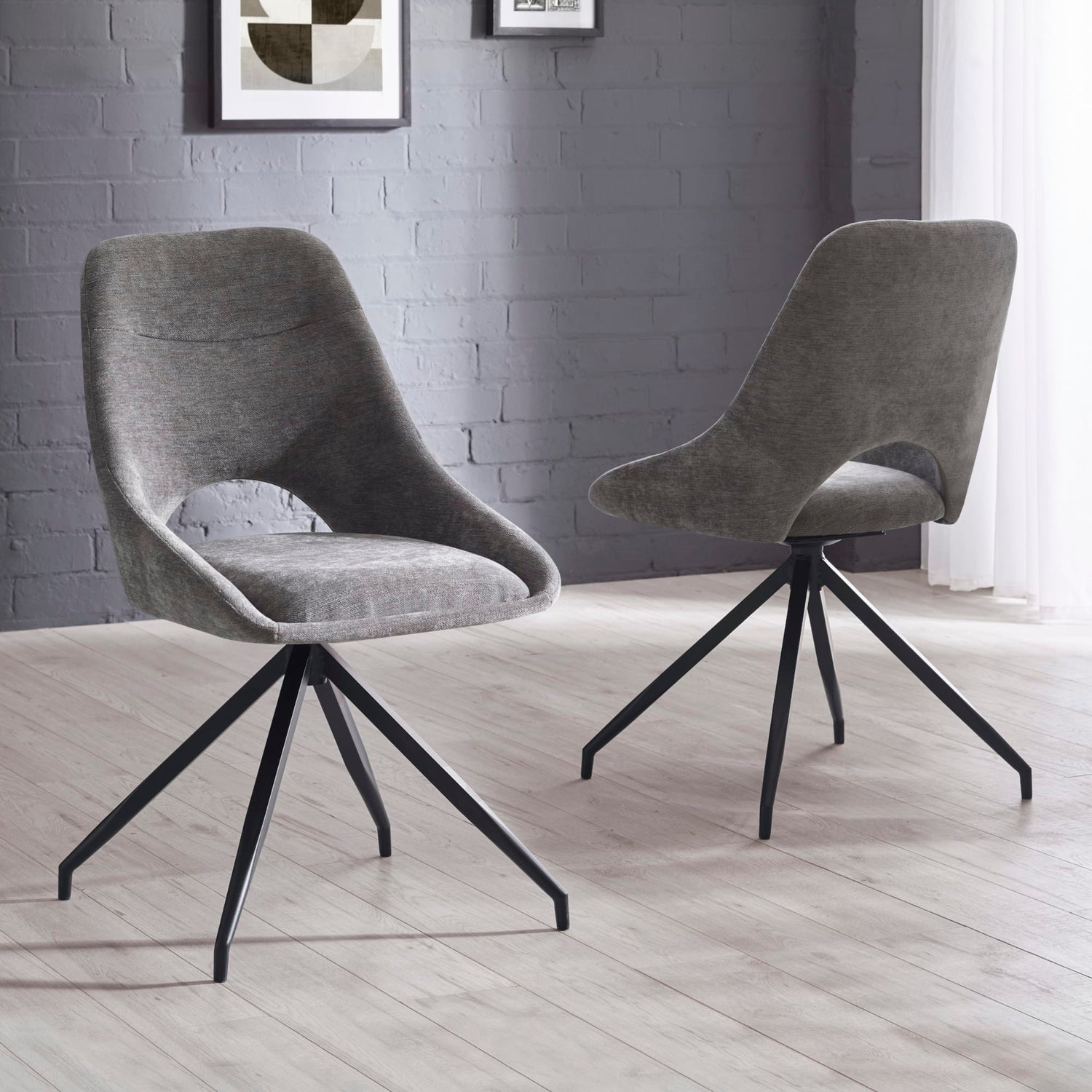 Two gray upholstered chairs with black metal legs on a light wooden floor against a gray brick wall.