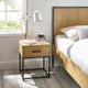 Wooden bedside table with lamp and cup next to a bed in a bright bedroom.