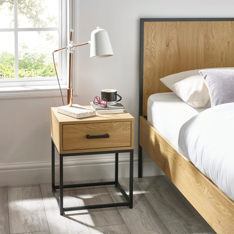 Wooden bedside table with lamp and cup next to a bed in a bright bedroom.