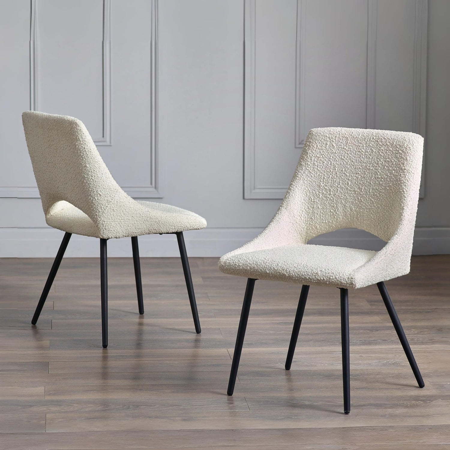 Two beige textured chairs with black legs on a wooden floor against a gray paneled wall.