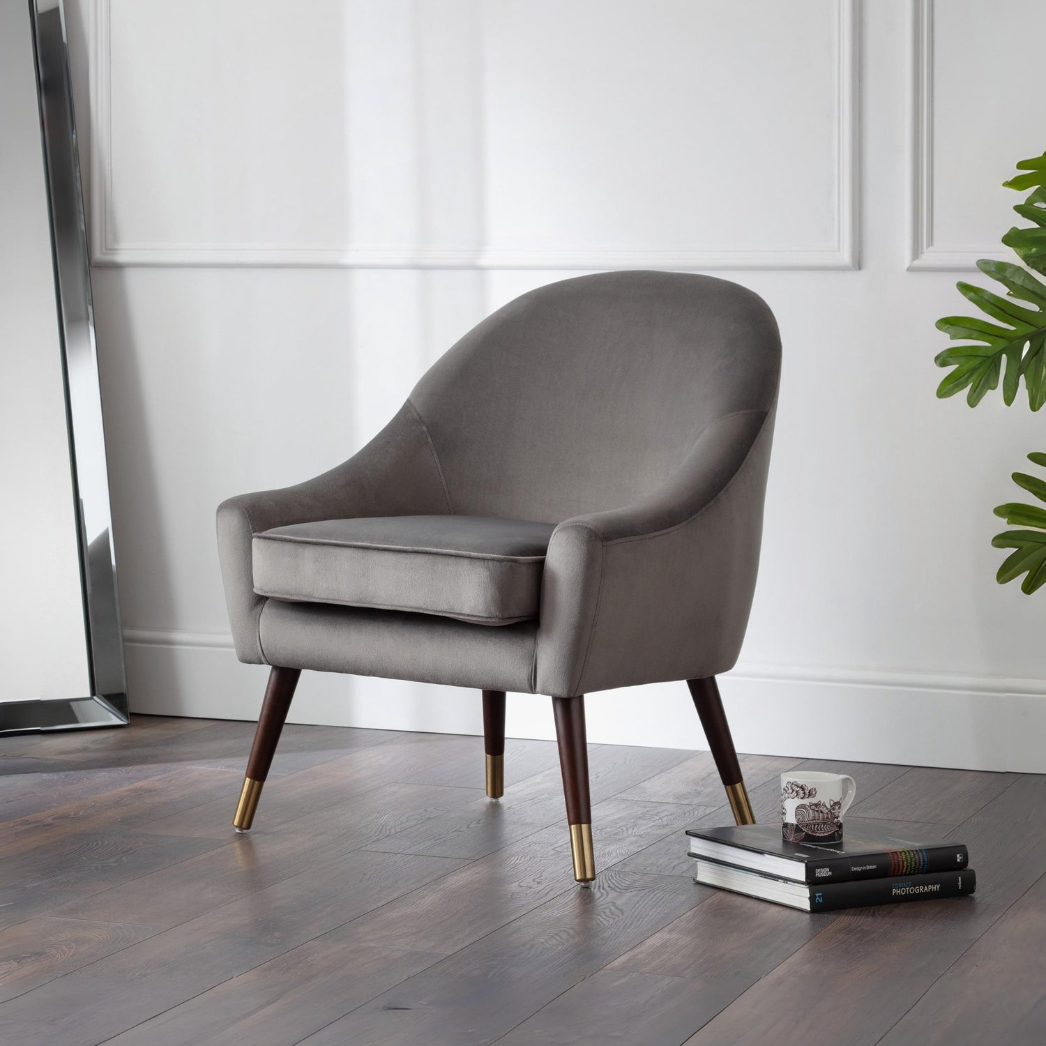 Gray armchair with wooden legs on a wooden floor, with books and a plant in the background.