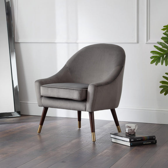 Gray armchair with wooden legs on a wooden floor, with books and a plant in the background.