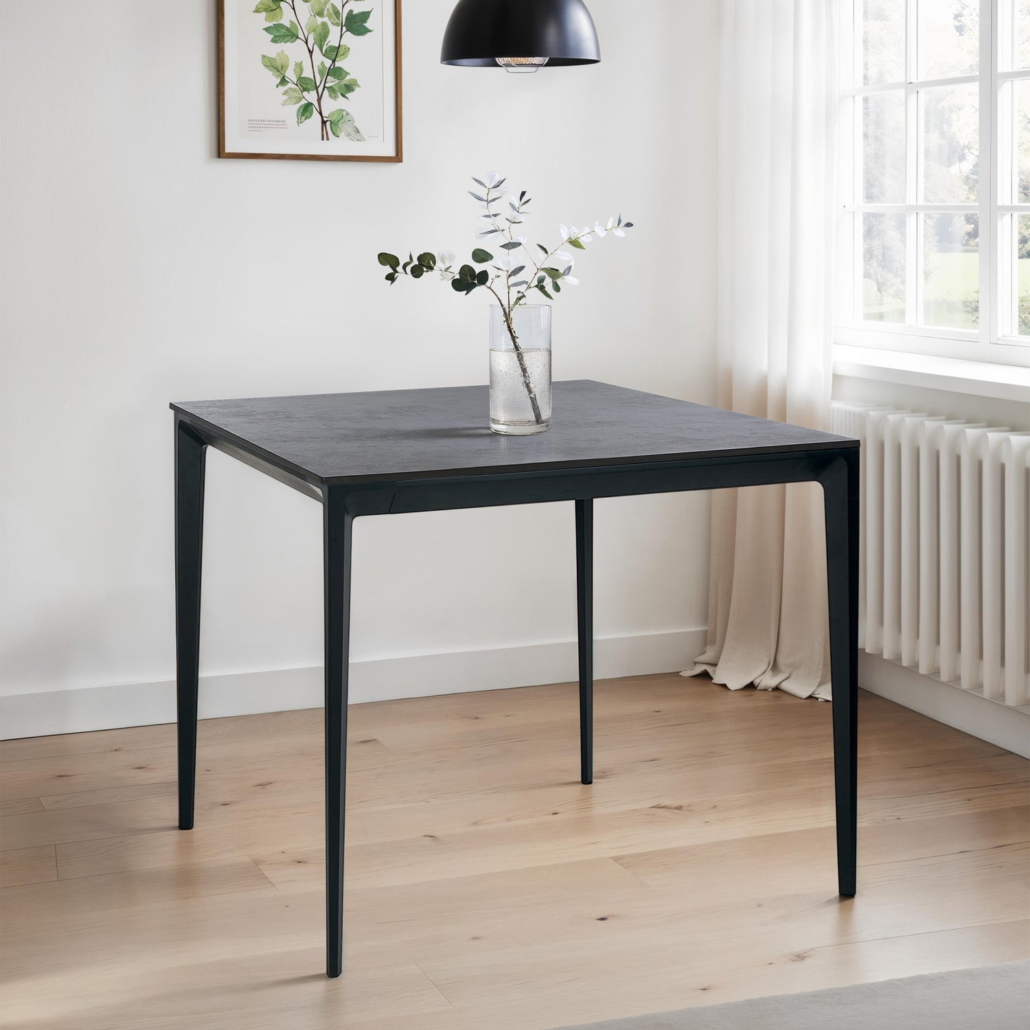 Black square table with a vase of flowers in a room with light wood flooring and a window.