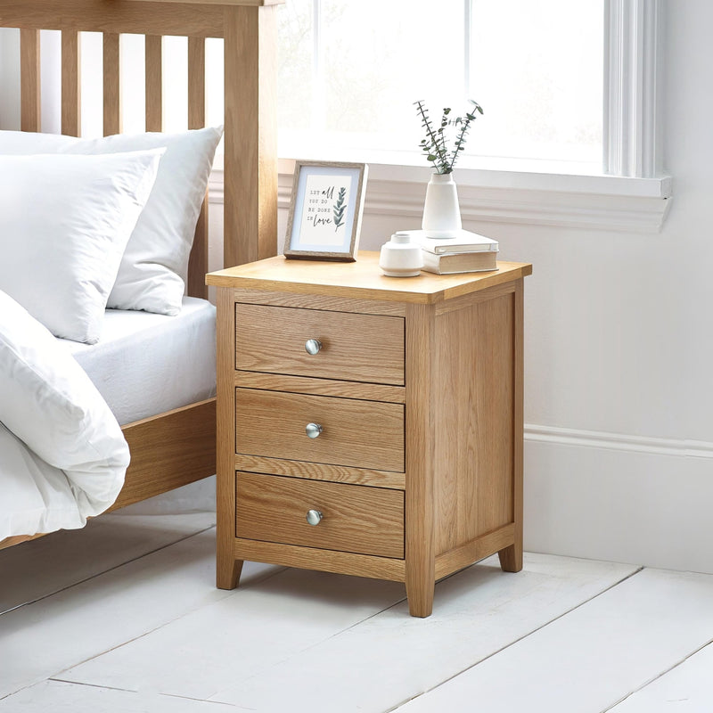Wooden nightstand with three drawers next to a bed in a bedroom setting.
