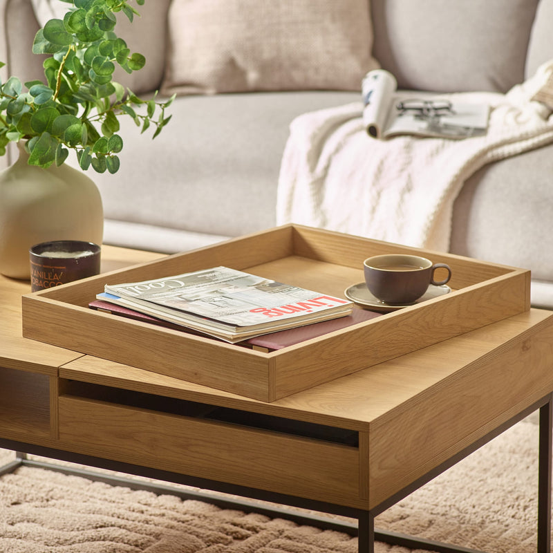 Wooden coffee table with a tray holding a cup, saucer, and books, with a plant and sofa in the background.