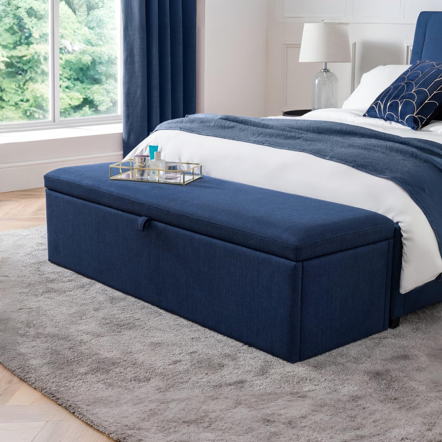 Navy blue ottoman at the end of a bed in a bedroom with a lamp and window in the background.
