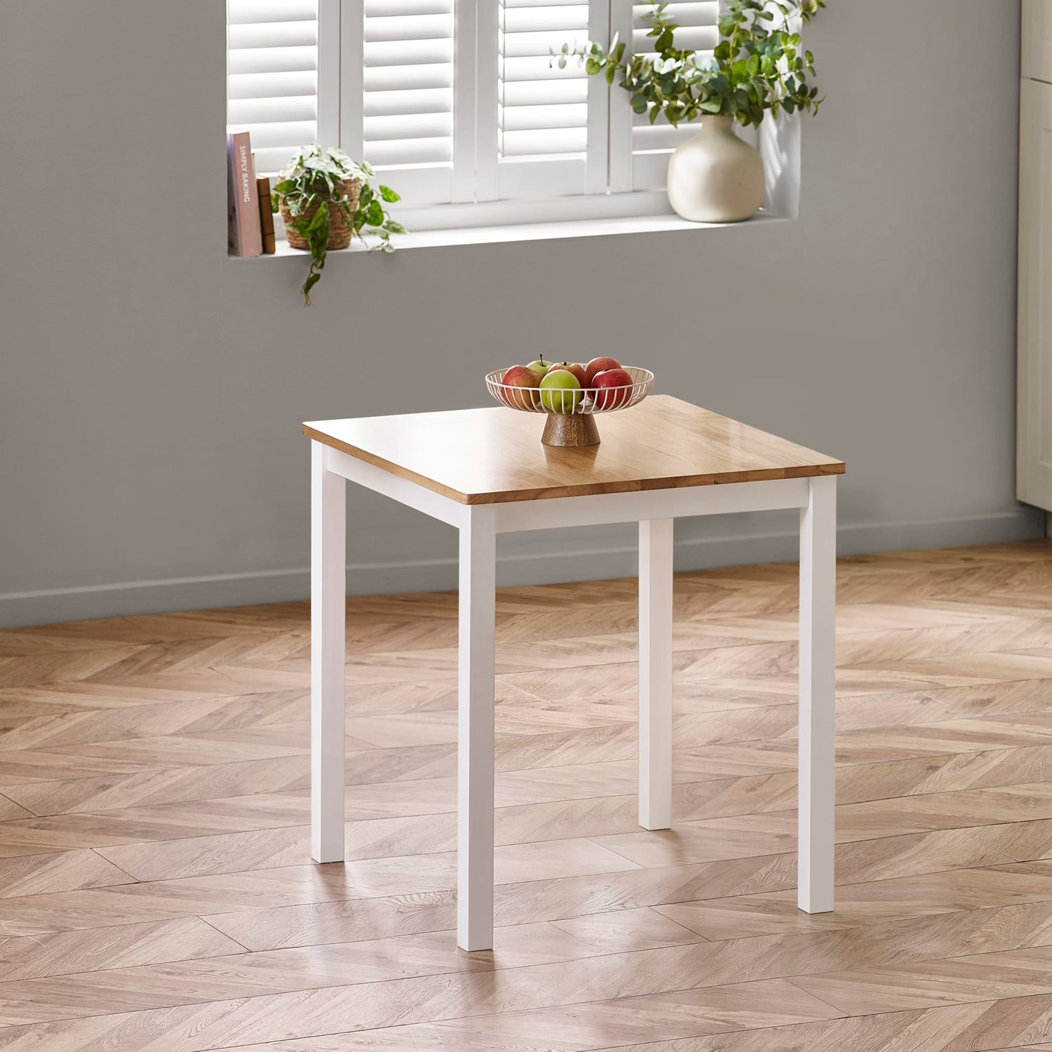 Square dining table with wooden top and white base in a room with a window and plants.