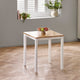 Square dining table with wooden top and white base in a room with a window and plants.