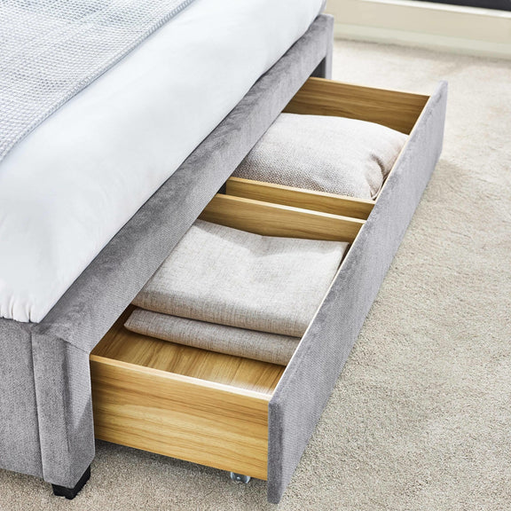 Gray bed frame with open wooden drawer containing folded bedding on a carpeted floor.