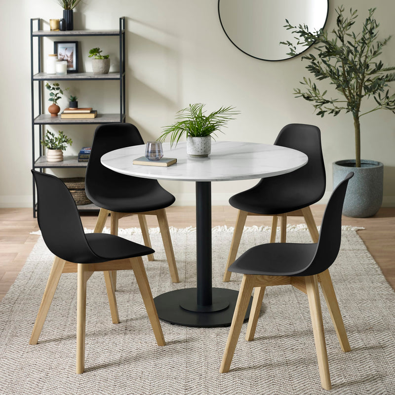 Dining area with a round table and black chairs on a rug, with plants and shelves in the background.
