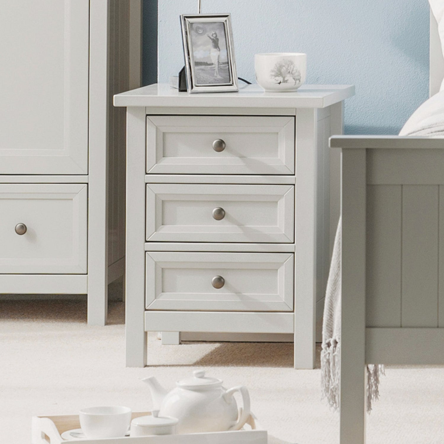 White nightstand with three drawers next to a bed in a bedroom setting.