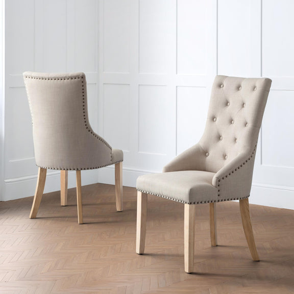 Two beige upholstered chairs with wooden legs on a wooden floor against a white paneled wall.