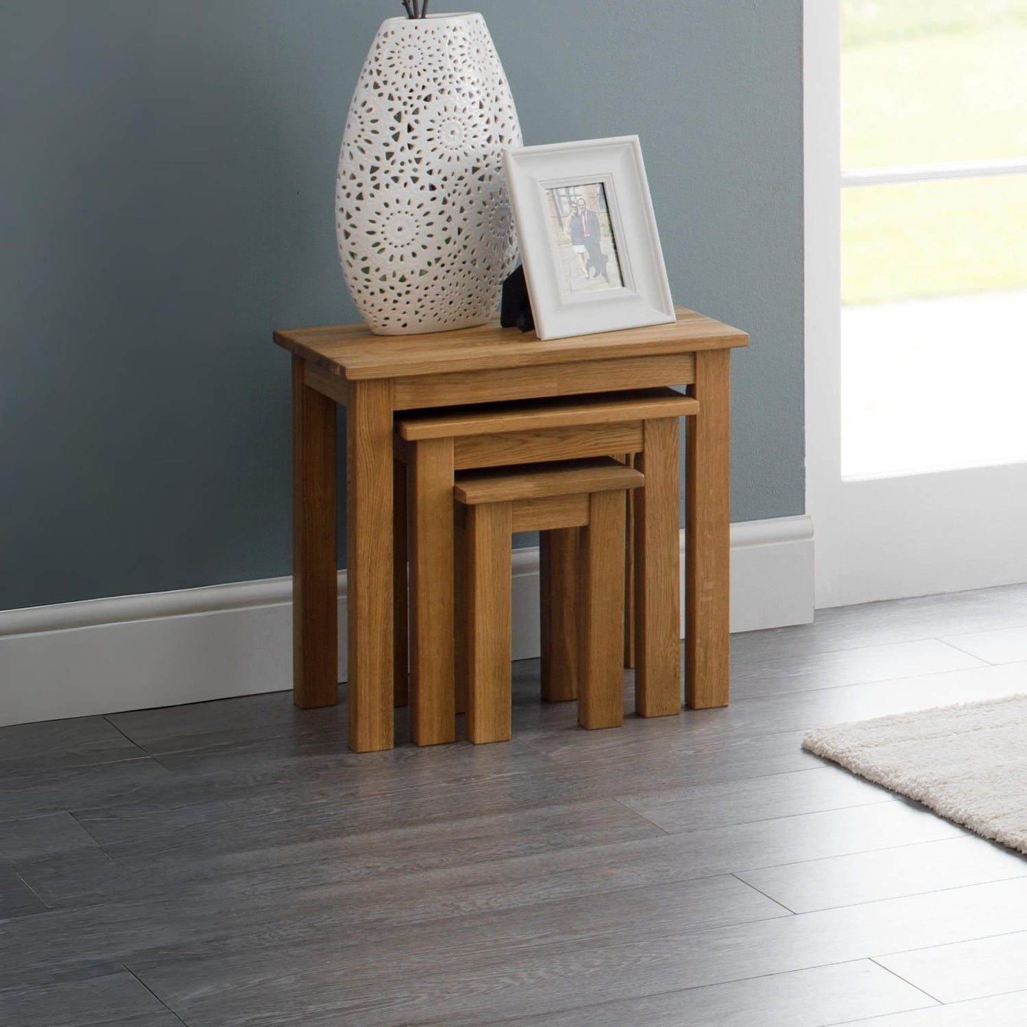 Set of three wooden nesting tables with a decorative vase and photo frame on a gray wall background.