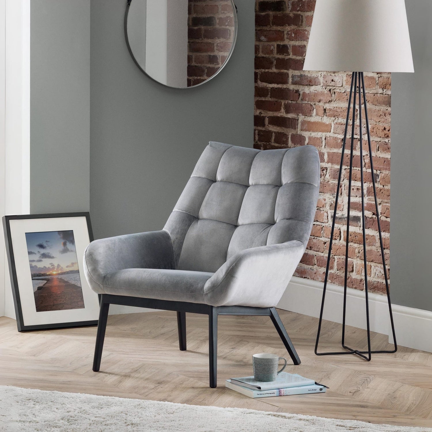 Gray armchair in a room with a brick wall, lamp, and framed picture.