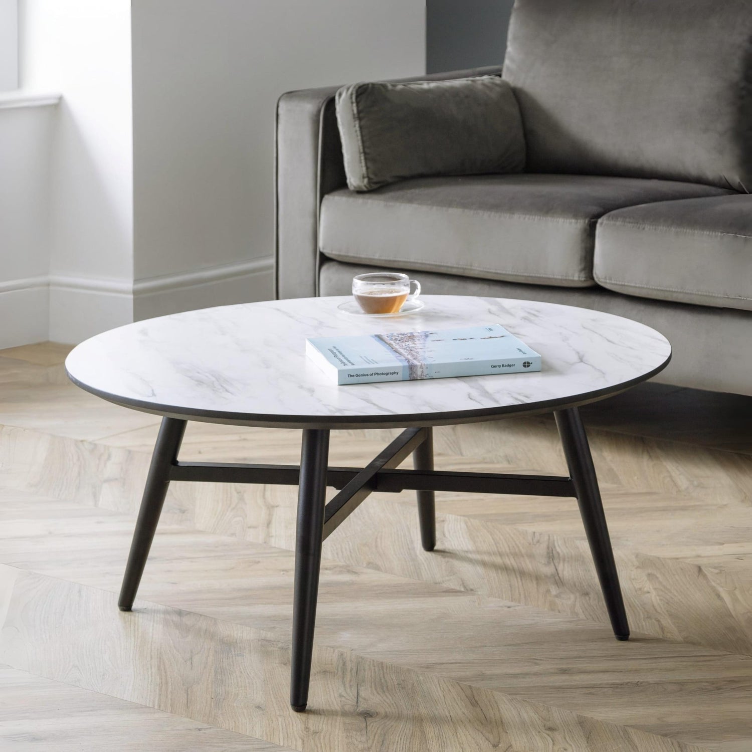 Round coffee table with a book and cup on it, placed in front of a gray sofa in a living room.