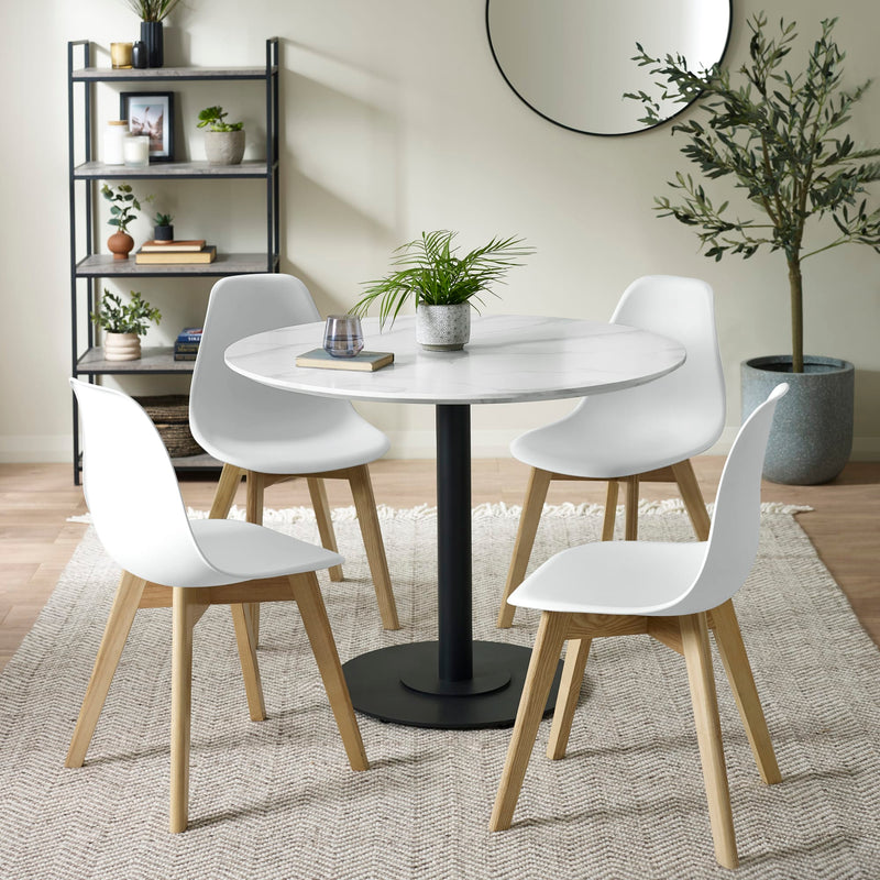 Dining area with a round table and white chairs on a rug, with a bookshelf and plants in the background.