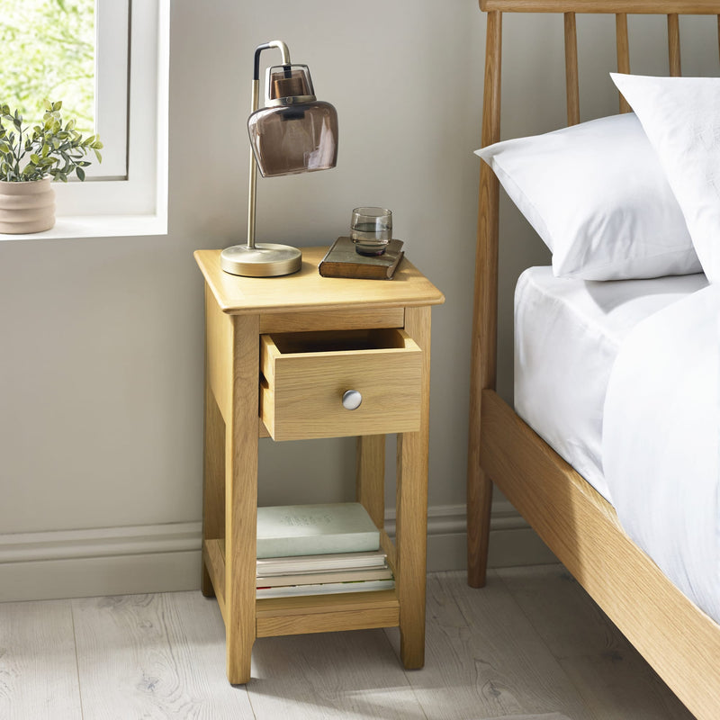 Wooden bedside table with lamp and books next to a bed in a bedroom.