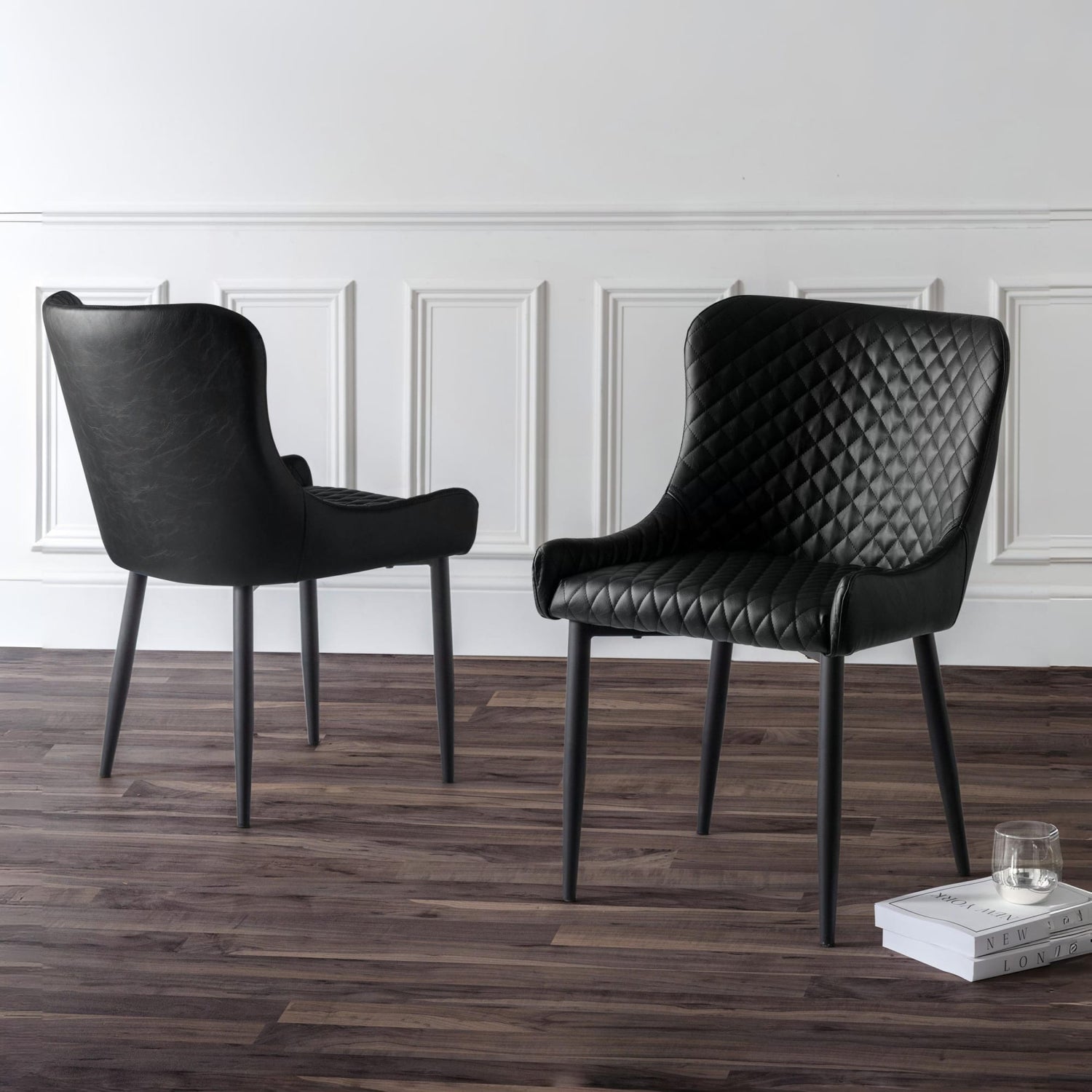 Two black chairs on a wooden floor with a white paneled wall in the background.