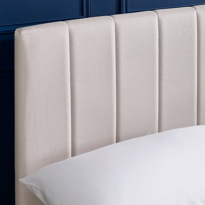Beige upholstered headboard with a pillow on a bed against a dark blue wall.