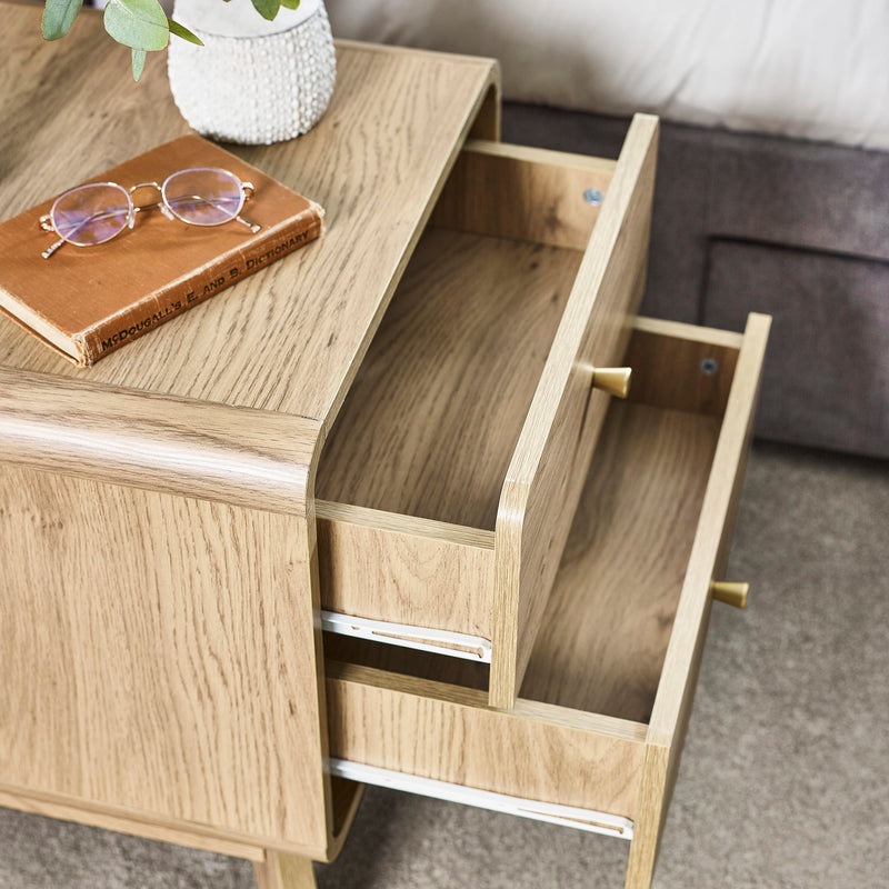 Wooden side table with an open drawer, glasses on a book, and a plant in the background.