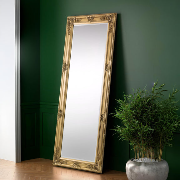 Gold-framed mirror leaning against a green wall with a plant on a wooden floor.