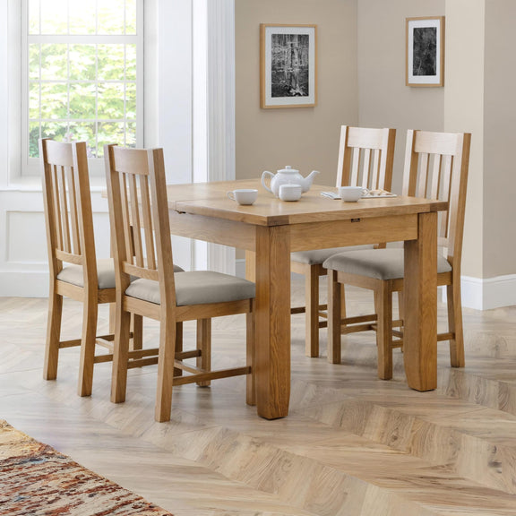Wooden dining table with four chairs in a bright room with large windows.