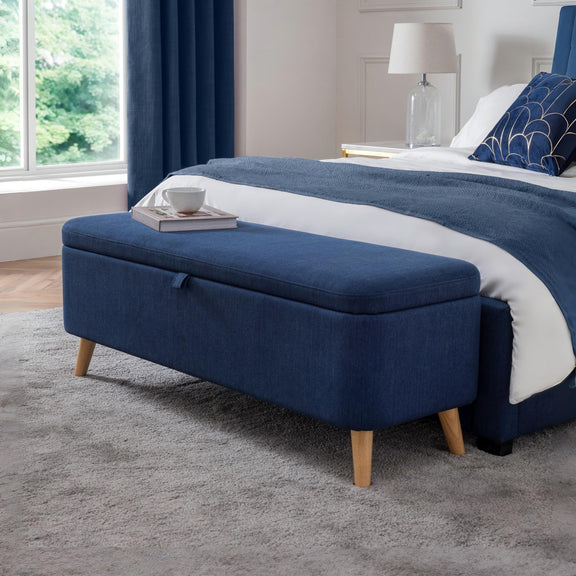 Blue ottoman with wooden legs in a bedroom setting.