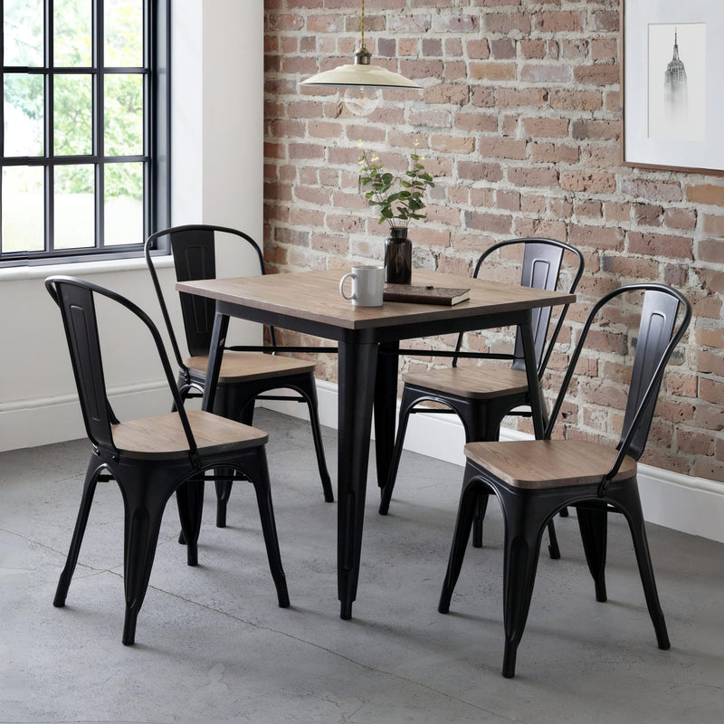 Dining area with a square table and black metal chairs against a brick wall.