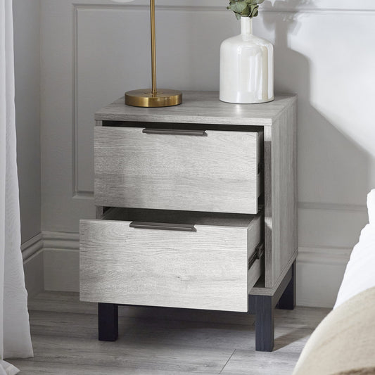 Wooden nightstand with two drawers, lamp, and vase against a neutral wall.