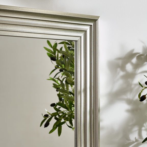 Silver-framed mirror with a reflection of green leaves on a white wall.