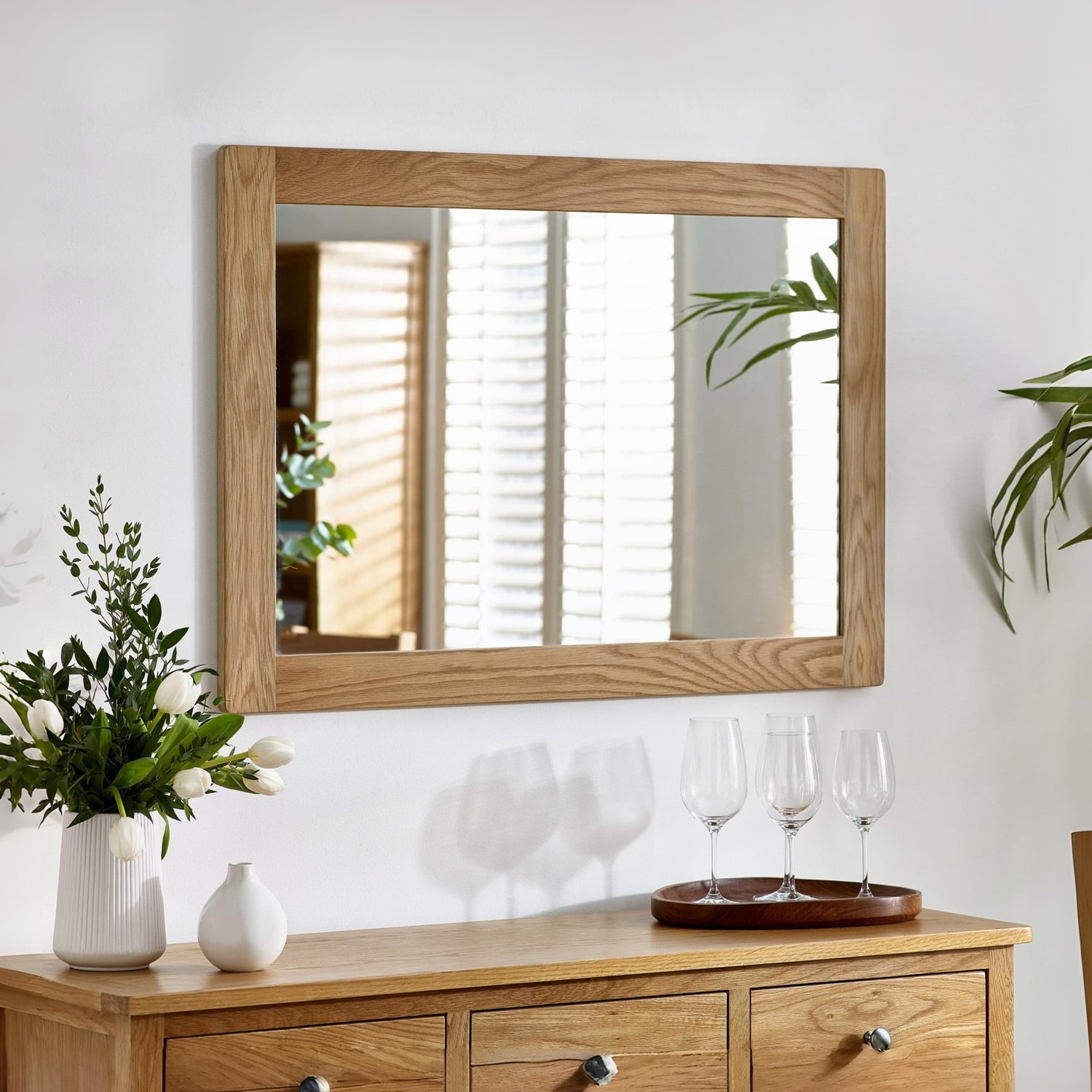 Wooden dresser with mirror, flowers, and glasses on a white wall.
