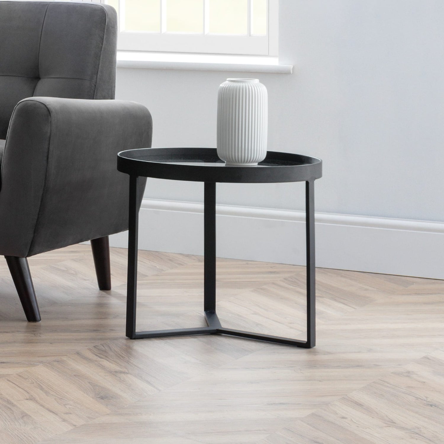 Black side table with a white vase next to a gray armchair in a room with wooden flooring and a window.