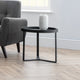 Black side table with a white vase next to a gray armchair in a room with wooden flooring and a window.