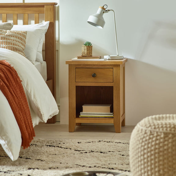 Wooden bedside table with lamp and books next to a bed with white bedding and orange throw.