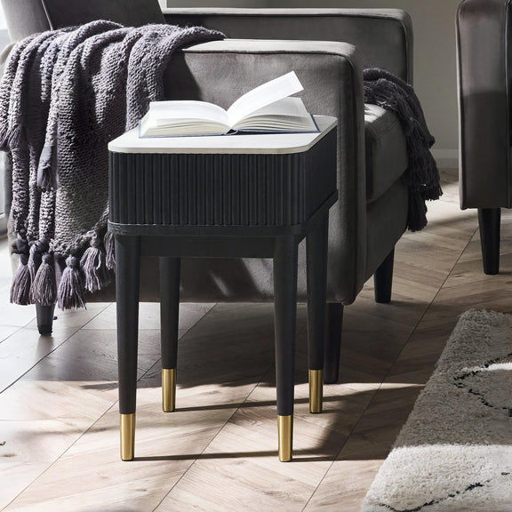 Black side table with gold legs next to a gray armchair with an open book on a wooden floor.