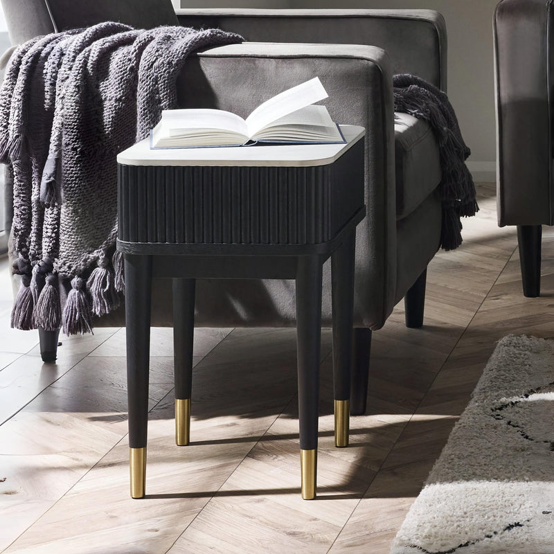 Black side table with gold legs next to a gray armchair with an open book on a wooden floor.