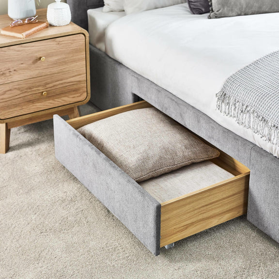 Gray upholstered bed with open drawer revealing a pillow, next to a wooden nightstand.