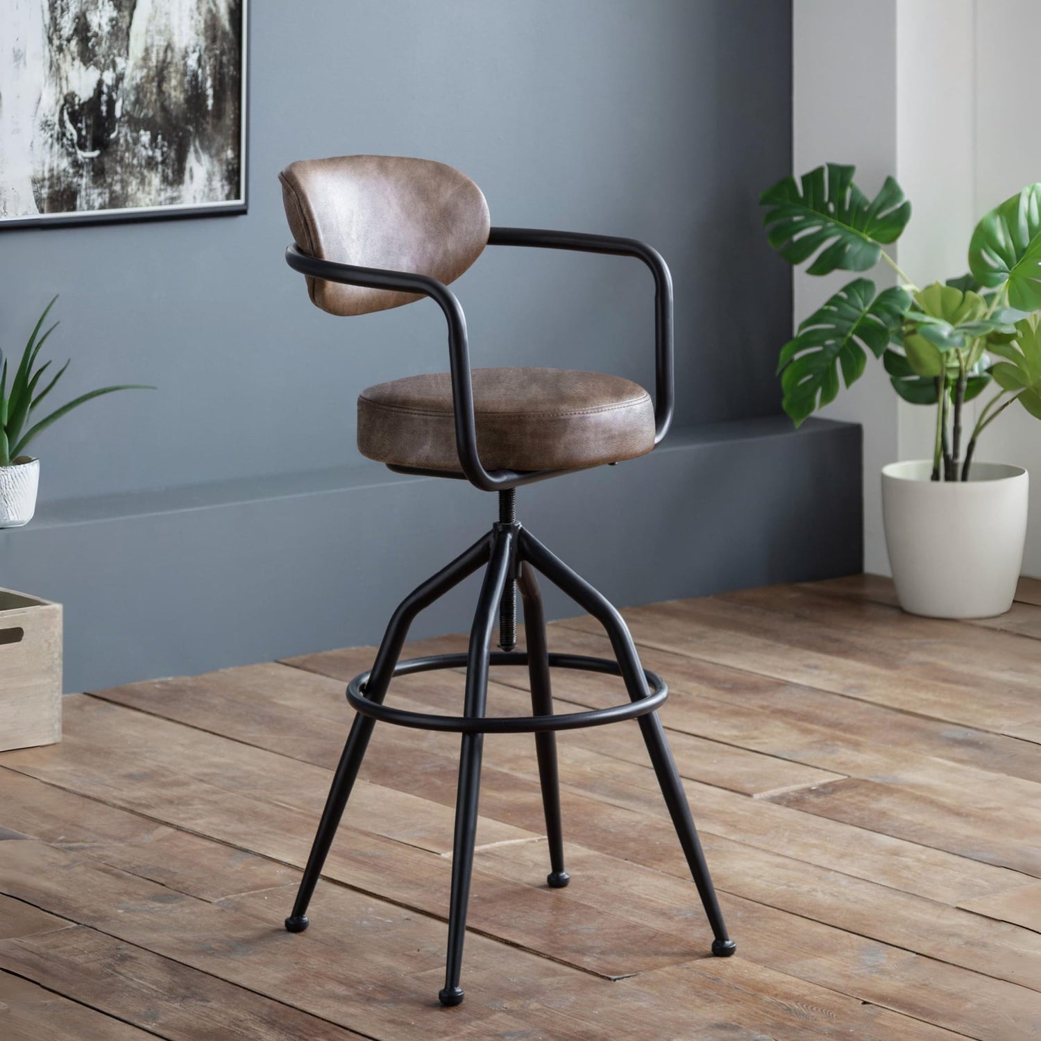 Brown bar stool with black metal frame in a room with plants and wooden floor.