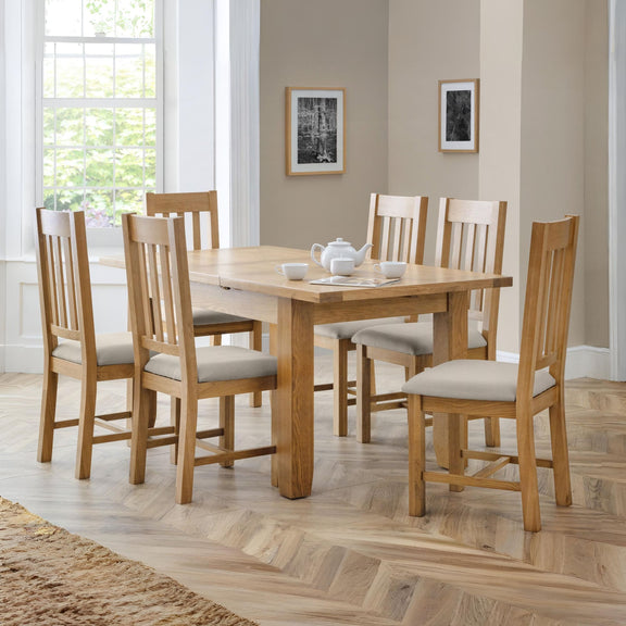 Wooden dining table with six chairs in a well-lit room with large windows.
