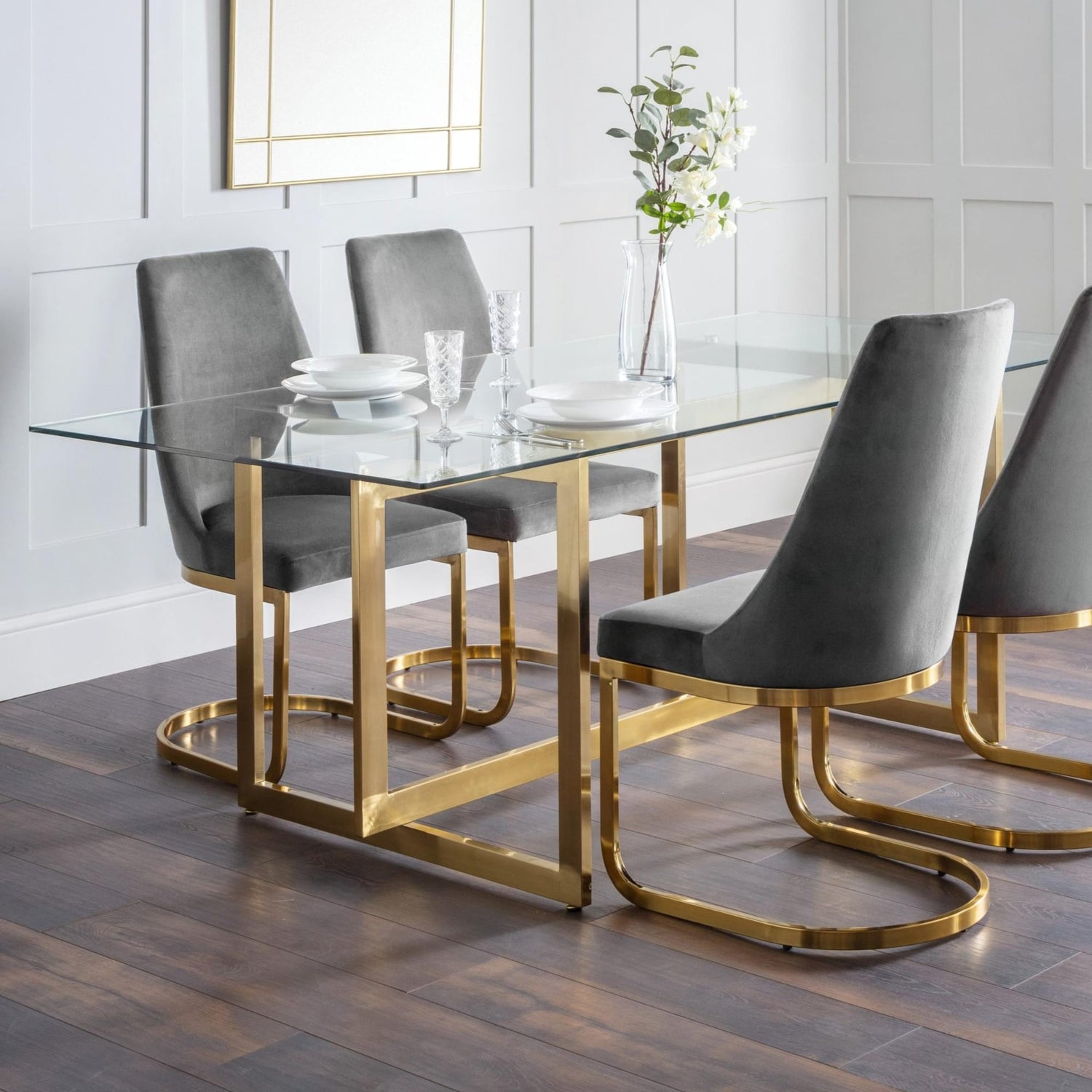 Dining table with glass top and gold legs accompanied by gray chairs in a modern setting.