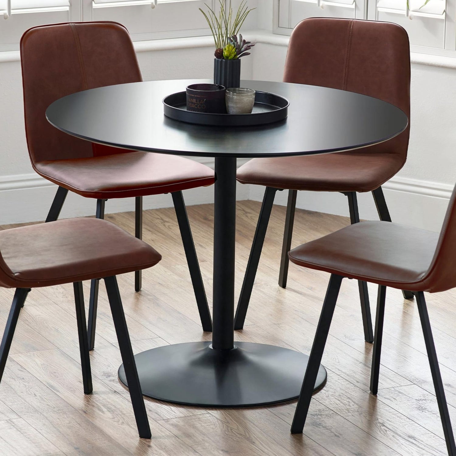 Round black dining table with four brown chairs in a room with large windows.