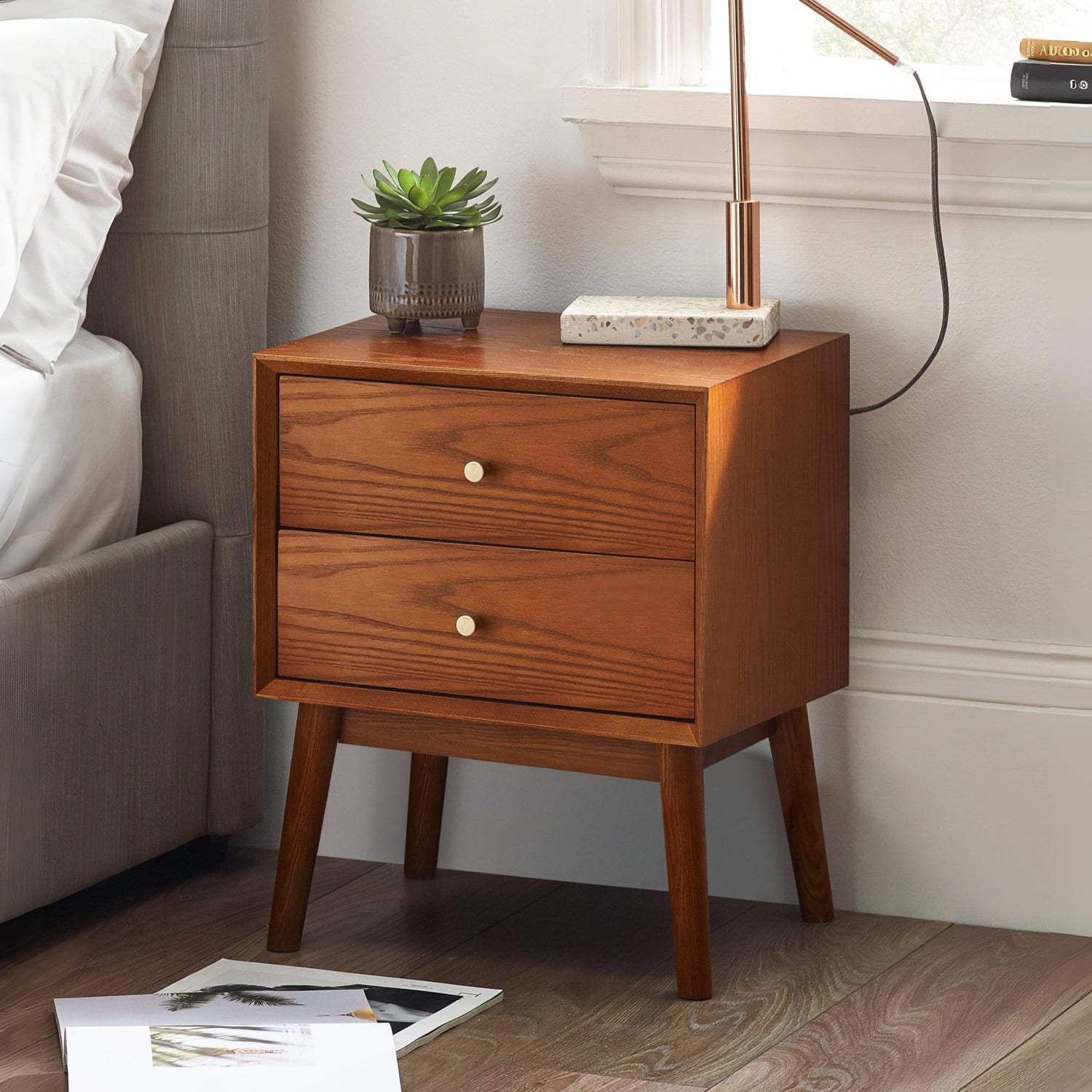 Wooden nightstand with two drawers, a plant, and a lamp in a bedroom setting.
