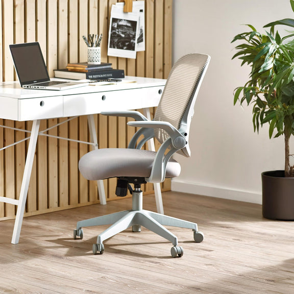 Gray office chair in front of a white desk with a laptop and books, in a room with wooden flooring and a plant.