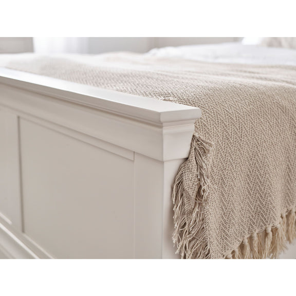 Beige throw blanket with fringes draped over a white wooden bed frame.