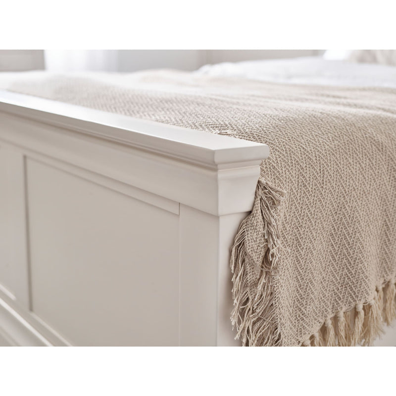 Beige throw blanket with fringes draped over a white wooden bed frame.