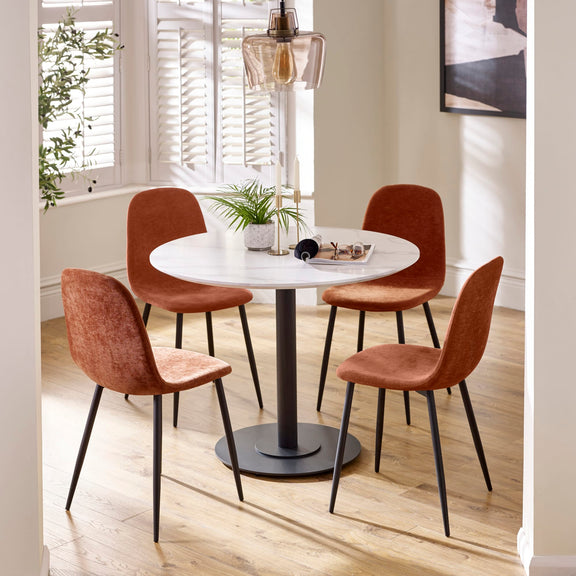 Round dining table with four brown chairs in a bright room with plants and a lamp.