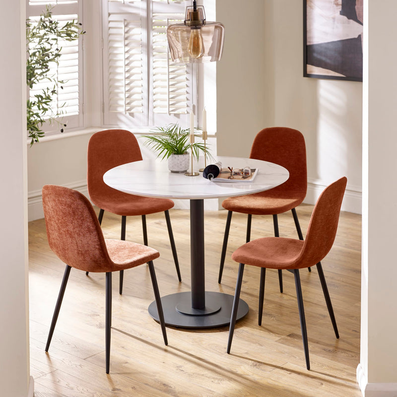 Round dining table with four brown chairs in a bright room with plants and a lamp.