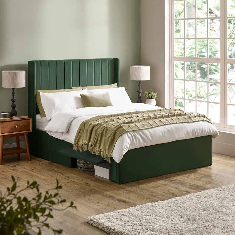 Green upholstered bed in a bedroom with wooden flooring and large windows.