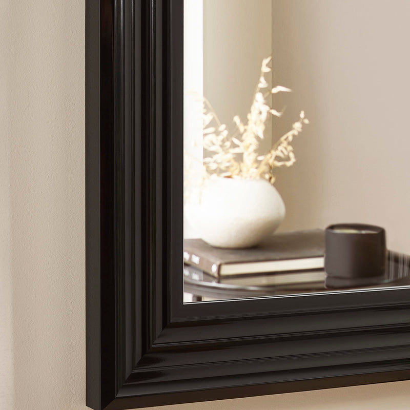 Black-framed mirror reflecting a decorative setup with a vase and candle.